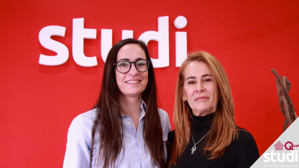 El nostre equip d'administració de finques: la Marta i l'Ana davant una pared vermella amb el logo d'Studi